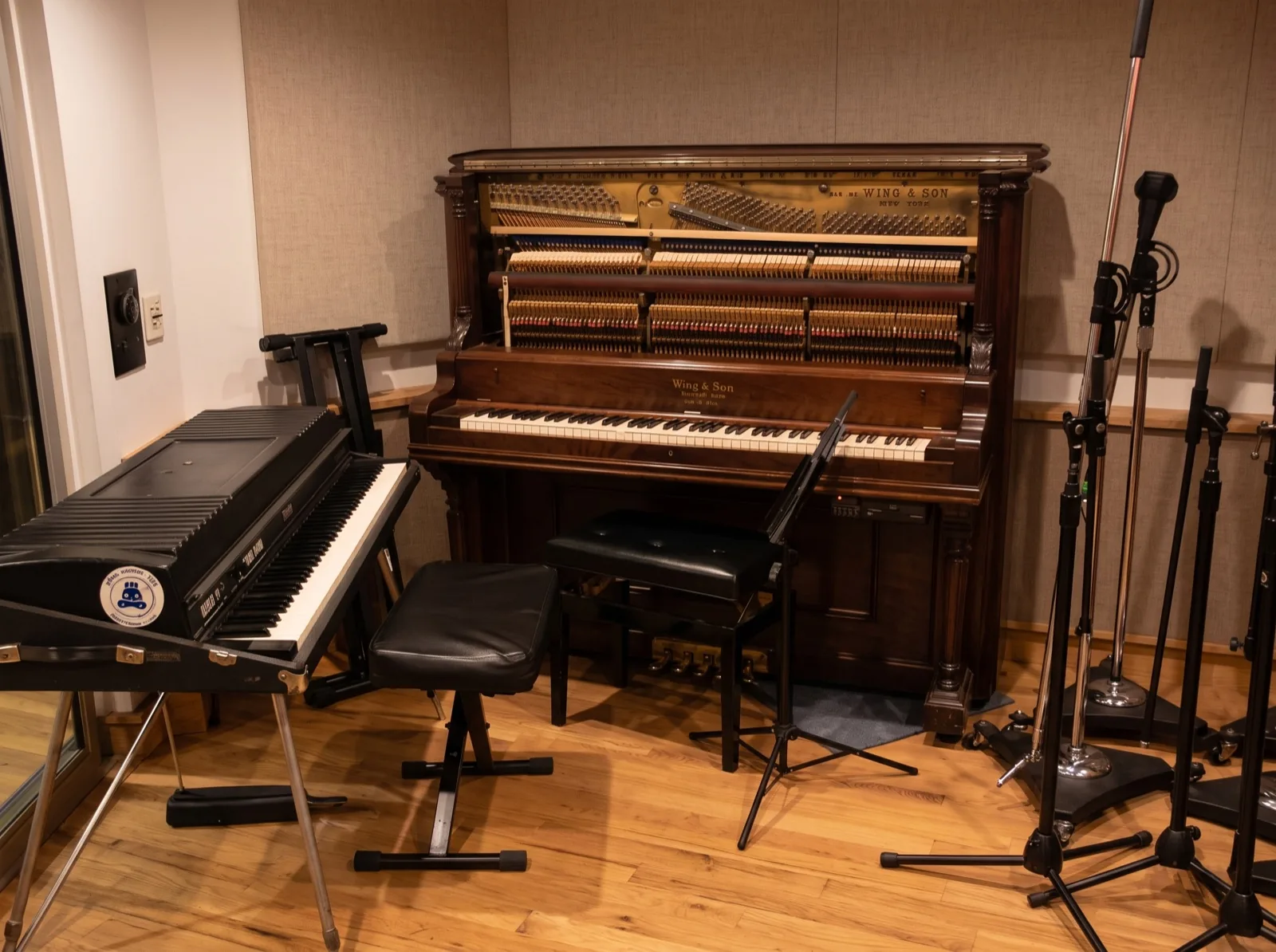 A Wing & Son upright piano with its front open, beside a vintage electric piano and a row of microphone stands